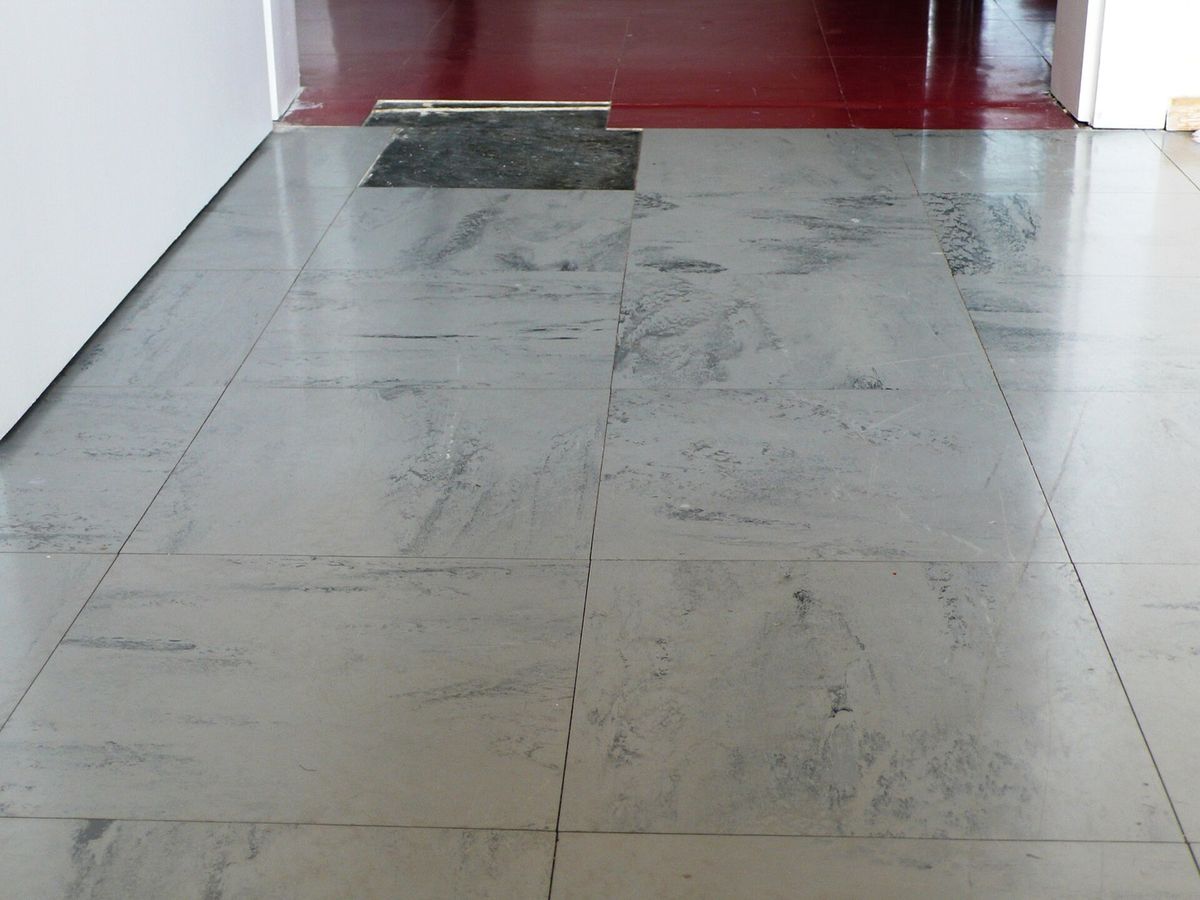 A floor of large pale grey vinyl tiles with dark mottling, one tile lifted to show the black bitumen bed beneath.